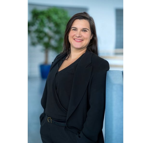 A woman with dark hair is smiling and standing indoors, wearing a black outfit and blazer. The background is softly blurred, showing a modern office space with a plant in the distance.