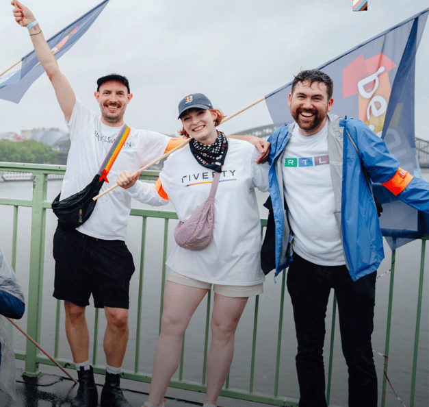 Three people standing on a bridge, smiling and posing together while holding pride flags. They are wearing casual clothes and T-shirts with Riverty and RTL logos, celebrating in a cheerful and inclusive atmosphere.
