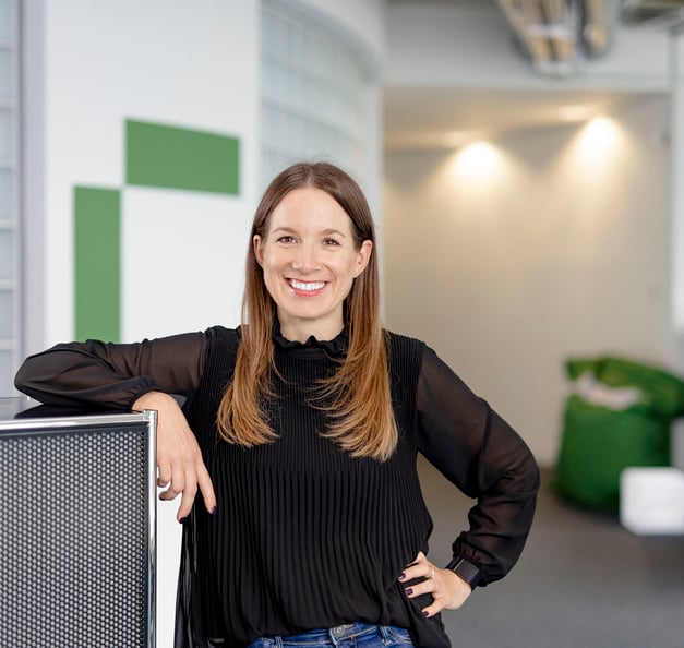 A woman with long brown hair is smiling and standing in a modern office space, leaning casually on a metal cabinet. She is wearing a black blouse and jeans, with soft lighting and green accents in the background.