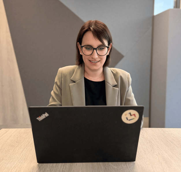 Eine Person mit dunklem, etwa schulterlangem Haar und dunkler Brille sitzt an einem hellen Holztisch und blickt auf einen geöffneten schwarzen Laptop. Die Person trägt einen beigefarbenen Blazer über einem schwarzen Oberteil sowie kleine Ohrstecker und eine feine Halskette. Auf der Rückseite des Laptops sind das „ThinkPad“-Logo und ein runder Aufkleber mit einem mehrfarbigen abstrakten Symbol zu sehen. Der Innenraum ist modern und zurückhaltend gestaltet. Im Hintergrund befinden sich große graue Wandflächen mit geometrischen Formen sowie rechts ein hoher heller Wandabschnitt und ein Fenster. Helles, weiches Licht fällt von rechts in den Raum.