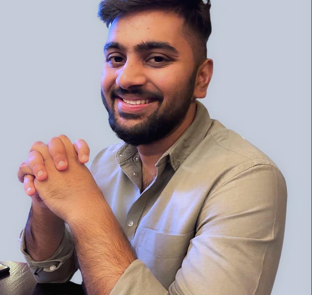 A man with short dark hair and a beard is smiling while sitting at a table with his hands clasped. He is wearing a beige shirt and facing the camera against a light blue background.