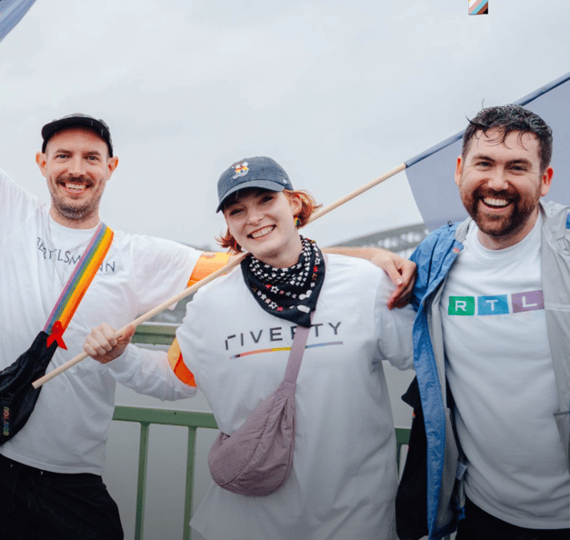 Three people standing on a bridge, smiling and posing together while holding pride flags. They are wearing casual clothes and T-shirts with Riverty and RTL logos, celebrating in a cheerful and inclusive atmosphere.
