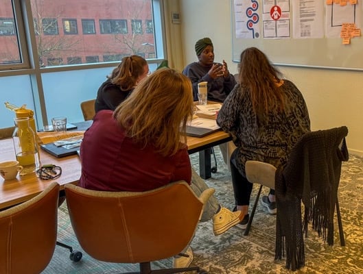 Workshopsessie bij Riverty waarbij deelnemers rond een tafel zitten en luisteren naar een ervaringsdeskundige van Stichting ONSbank, met notities en posters over jongeren en financiën zichtbaar aan de muur in een vergaderruimte.