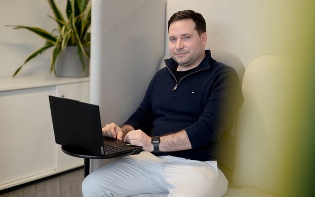 A man sits in a cushioned booth working on a laptop placed on a small table. He wears a dark sweater and light-colored pants, with a watch on his wrist, and looks toward the camera. A potted plant is visible on a cabinet in the background.