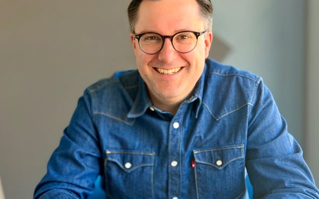 The image shows a portrait photo of Florian Schipperges. The person is visible from the shoulders up and looks directly into the camera. Florian smiles warmly, creating an open and approachable impression. The hair is short and medium brown. Florian wears simple, dark rounded glasses.  Florian is dressed in a dark blue denim shirt that fits casually. The background is light and slightly blurred, with no recognizable objects or text, keeping the focus on the face and upper body. No assistive devices, brand logos, or other people are visible in the image.