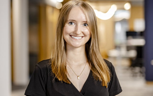 Portrait of a young professional woman with long blonde hair, smiling in an office environment, wearing a black v-neck top and a necklace with a letter pendant.