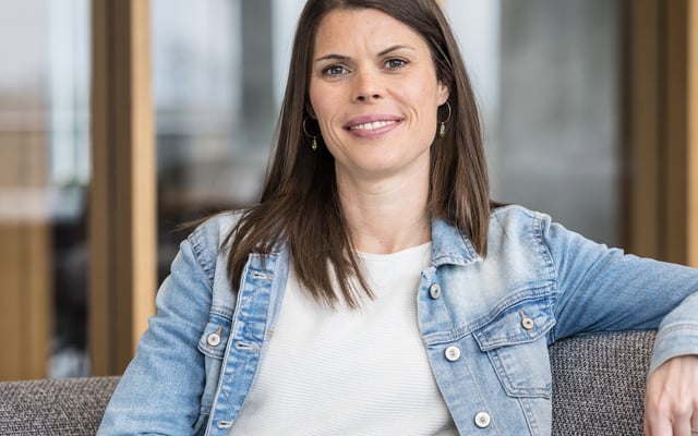 Eine Person mit schulterlangem, braunem Haar steht auf einem hellen Fliesenboden vor einem Gebäude mit einer Glasfront. Im Hintergrund ist in grüner Schrift der Text „RIVERTY LOUNGE" auf der Glasscheibe zu lesen, was auf eine Lounge oder einen Aufenthaltsbereich hinweist. Durch die Glasfront sind Pflanzen sowie Möbel zu erkennen. Die Person trägt ein weißes, kurzärmeliges T-Shirt, eine olivgrüne Hose und hält eine hellblaue Jeansjacke über den Arm. Über der Schulter trägt sie eine olivgrüne Umhängetasche. Am Handgelenk ist ein schwarzes Armband sichtbar. Die Person hält ein schwarzes Smartphone ans Ohr und telefoniert. Der Gesichtsausdruck ist offen und freundlich, die Person lächelt breit und schaut leicht nach links, weg von der Kamera. Die Körperhaltung wirkt entspannt und zugewandt.
