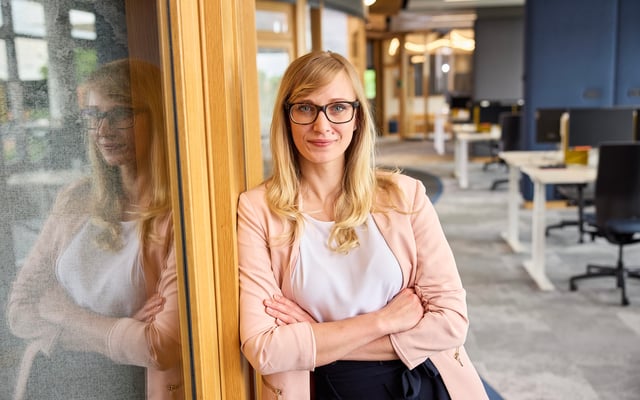 A blonde woman with glasses, wearing a light pink blazer and white top, leans with folded arms against a wooden doorframe, with her reflection visible on a adjacent glass panel in a modern office space.