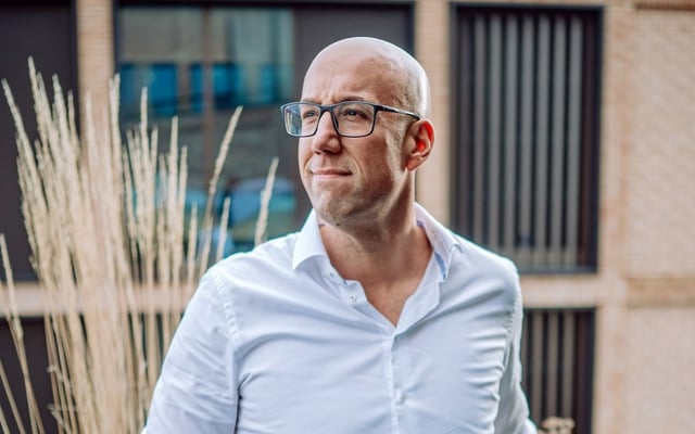 Kale man met een bril en een wit overhemd die buiten staat en bedachtzaam opzij kijkt, met een modern gebouw op de achtergrond
