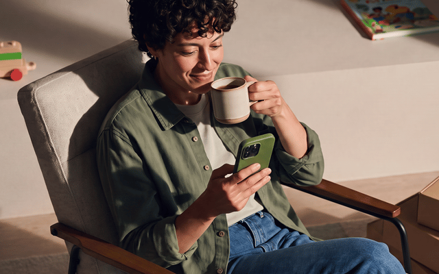 Person sitting relaxed in an armchair, looking at a smartphone and holding a mug.
