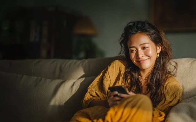 Lächelnde junge Frau sitzt auf einem beigen Sofa, trägt ein senfgelbes Outfit, hält ein Smartphone in der Hand und genießt einen entspannten Moment in einem gemütlich beleuchteten Wohnzimmer.