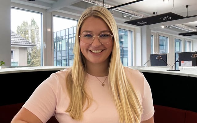 Portrait of a young professional woman with long blonde hair, smiling in an office environment, wearing a black v-neck top and a necklace with a letter pendant.