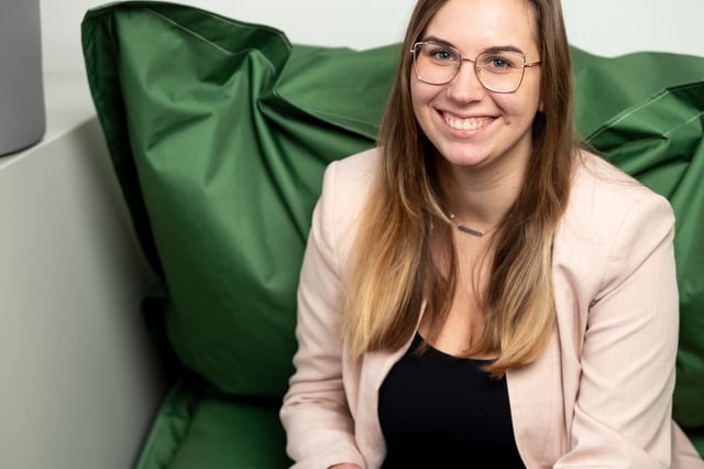 Woman with long hair and glasses wearing a light blazer and dark top, sitting on a green beanbag chair and smiling