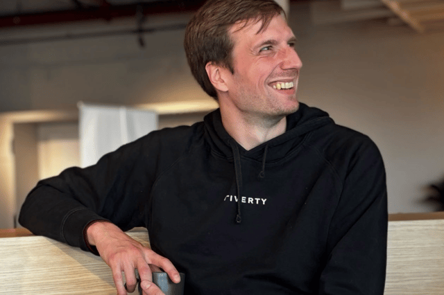 Max im schwarzen Riverty-Hoodie sitzt entspannt an einem hellen Holztisch, hält eine Tasse in der Hand und blickt zur Seite.