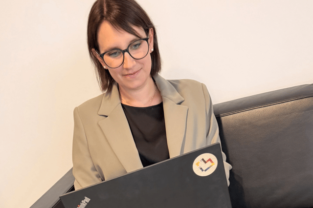 Eine Person mit dunklem, etwa schulterlangem Haar und dunkler Brille sitzt auf einem schwarzen Ledersofa vor einer hellen, einfarbigen Wand. Die Person trägt einen beigefarbenen Blazer über einem schwarzen Oberteil und blickt nach unten auf einen geöffneten schwarzen Laptop. Auf dem Laptop ist links das „ThinkPad“-Logo zu sehen. Oben rechts befindet sich ein runder Aufkleber mit einem mehrfarbigen, abstrakten Symbol. Die Aufnahme ist hell und in warmen, neutralen Farbtönen gehalten.