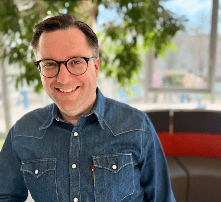 uf dem Foto ist eine Person zu sehen, die direkt in die Kamera schaut und herzlich lächelt. Die Person trägt eine runde, schwarze Hornbrille und ein mittelblau gefärbtes Jeanshemd der Marke Levi's, erkennbar am roten Markenzeichen auf der Brusttasche. Das Hemd hat zwei aufgesetzte Brusttaschen mit weißen Knöpfen und sichtbaren gelben Nähten. Die Person hat kurzes, dunkelbraunes Haar mit leichten grauen Akzenten an den Seiten. Der Gesichtsausdruck wirkt offen, einladend und positiv. Im Hintergrund ist ein heller, moderner Innenraum zu erkennen. Links im Bild sind große Glasfenster sichtbar, durch die Grünpflanzen und ein blauer Himmel zu sehen sind. Rechts hinter der Person befindet sich eine geschwungene Sitzbank mit einem dunkelbraunen und einem roten Polsterabschnitt. Über der Person hängen Äste mit grünen Blättern, vermutlich eines Zimmerbaums. Der Hintergrund ist leicht unscharf (Bokeh-Effekt), wodurch die Person klar im Fokus steht. Es sind keine Textelemente, Bildschirme, Symbole oder technische Geräte im Bild sichtbar.