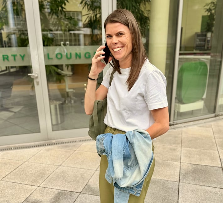 The image shows a person standing outdoors in front of a modern building while talking on the phone. The person has long, straight, dark brown hair worn open. Their facial expression is very cheerful and open: they are laughing broadly into the camera, conveying energy and a positive mood. The person is wearing a plain white t-shirt and olive green trousers. A light blue denim jacket is held loosely in one hand rather than being worn. An olive green shoulder bag is visible over one shoulder. A black bracelet is visible on the wrist. In one hand, the person is holding a dark smartphone pressed to their ear. In the background, a glazed building facade is visible with green lettering, of which the words "RIVERTY" and "LOUNGE" are legible. Through the glass panels, plants and seating furniture are visible inside the building. The ground consists of light stone tiles. The surroundings appear urban and publicly accessible.