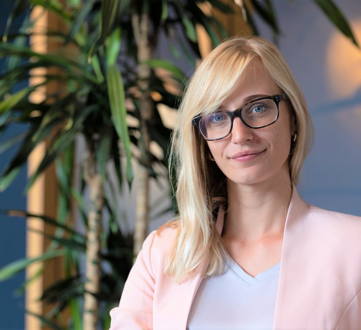 Bild von Regina im Büro, wie sie in die Kamera lächelt, im Hintergrund eine Zimmerpflanze