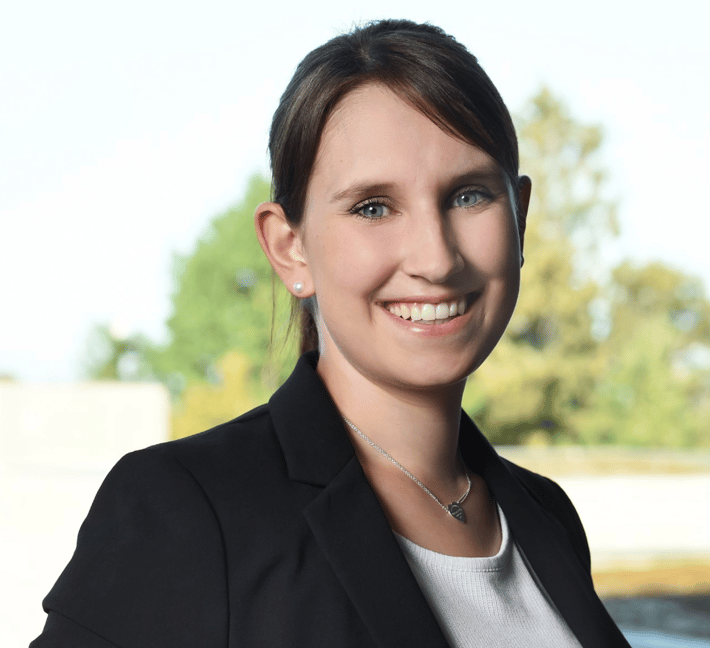 Das Bild zeigt ein professionell aufgenommenes Porträt einer Person mit dunklem, zurückgebundenem Haar. Die Person trägt einen schwarzen Blazer über einem hellgrauen Oberteil sowie kleine runde Ohrstecker und eine feine Halskette mit einem kleinen herzförmigen Anhänger. Die Person blickt direkt in die Kamera; die Zähne sind sichtbar. Der Oberkörper ist leicht seitlich ausgerichtet. Im Hintergrund befindet sich eine große Fensterfläche. Dahinter sind grüne und gelbliche Bäume sowie Teile eines Gebäudes unscharf zu erkennen. Helles Tageslicht und der stark verschwommene Hintergrund heben die Person deutlich hervor.