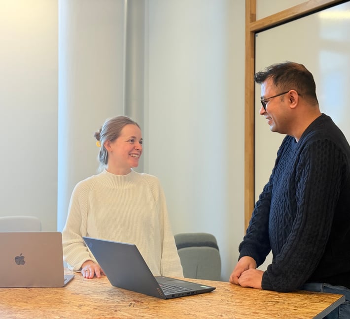 Zwei Personen stehen an einem Holztisch in einem hellen Besprechungsraum, lächeln und unterhalten sich miteinander. Vor ihnen steht jeweils ein offener Laptop, und an der Wand hinter ihnen ist ein Whiteboard zu sehen.
