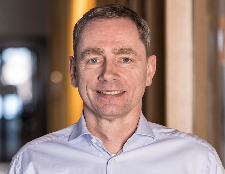 Portret van een middelbare man in een lichtblauwe overhemd die glimlachend in de camera kijkt in een zacht verlichte kantooromgeving.