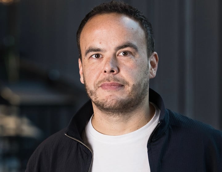 Professional portrait of a man in casual business attire standing in a modern office environment with soft lighting and a blurred background.