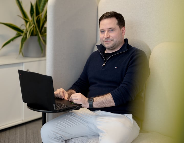 A person sits in a high-backed, cushioned office booth, using a black laptop placed on a small round table. They are wearing a dark navy zip-collar sweater, light-colored pants, and a smartwatch. A potted plant with long green leaves sits on a cabinet in the background, suggesting a modern, quiet workspace designed for focused work.