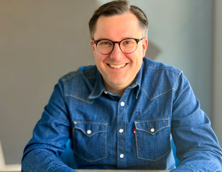 Das Bild zeigt ein Porträtfoto von Florian Schipperges. Die Person ist von den Schultern aufwärts zu sehen und blickt direkt in die Kamera. Florian lächelt freundlich, was einen offenen und nahbaren Eindruck vermittelt. Die Haare sind kurz und mittelbraun. Florian trägt eine schlichte, dunkle Brille mit runden Gläsern.  Florian hat ein dunkelblaues Jeanshemd an, das locker sitzt und eher lässig wirkt. Der Hintergrund ist hell und leicht unscharf, ohne erkennbare Gegenstände oder Texte, sodass der Fokus auf Gesicht und Oberkörper liegt. Es sind keine technischen Hilfsmittel, Markenlogos oder weiteren Personen im Bild zu sehen.