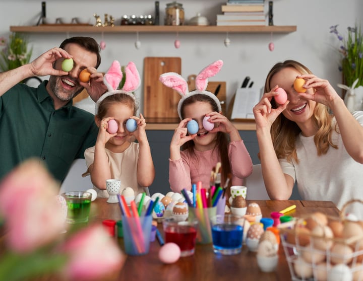 Eine glückliche Familie bemalt Ostereier und hält sich die bunten Ostereier als Augen vor ihre Gesichter