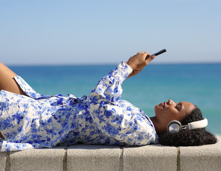 Frau liegt am Strand und hört mit Kopfhörern einen Finanzpodcast