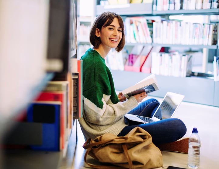 Eine lächelnde Studentin sitzt in einer Bibliothek auf dem Boden, liest ein Buch und arbeitet an ihrem Laptop.
