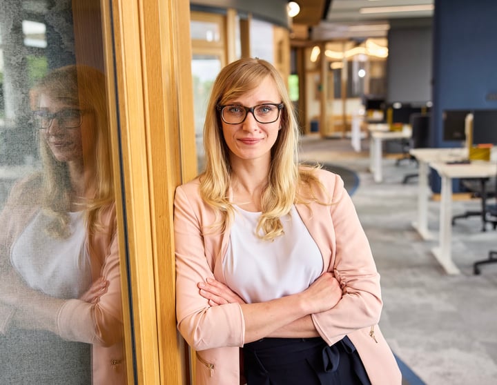 A blonde woman with glasses, wearing a light pink blazer and white top, leans with folded arms against a wooden doorframe, with her reflection visible on a adjacent glass panel in a modern office space.