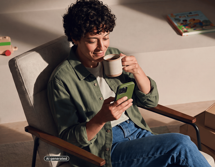 Person sitting relaxed in an armchair, looking at a smartphone and holding a mug.