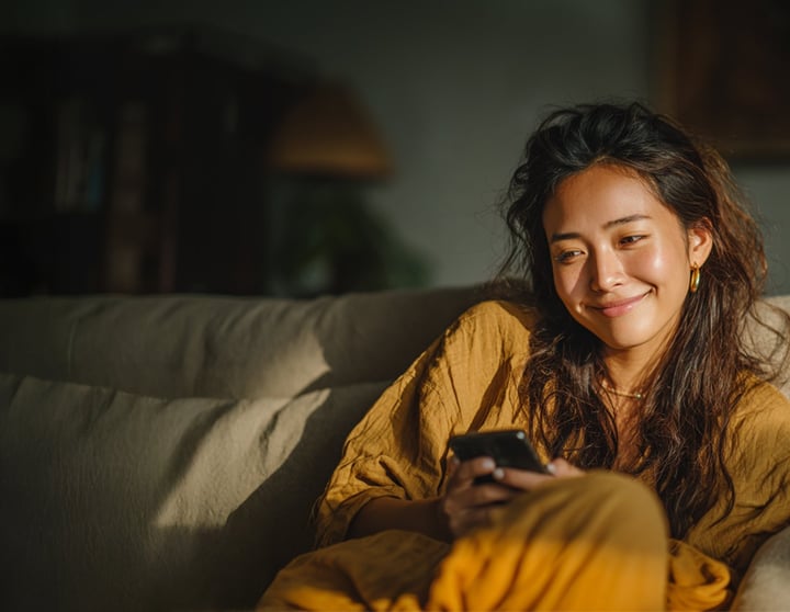 Lächelnde junge Frau sitzt auf einem beigen Sofa, trägt ein senfgelbes Outfit, hält ein Smartphone in der Hand und genießt einen entspannten Moment in einem gemütlich beleuchteten Wohnzimmer.