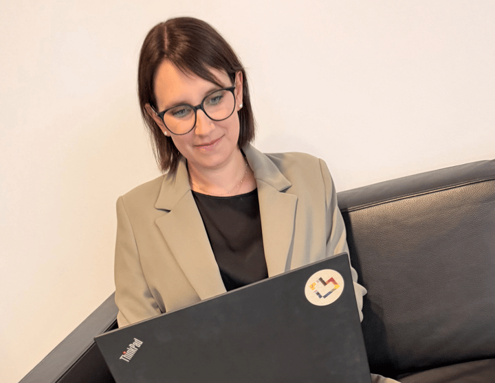 Eine Person mit dunklem, etwa schulterlangem Haar und dunkler Brille sitzt auf einem schwarzen Ledersofa vor einer hellen, einfarbigen Wand. Die Person trägt einen beigefarbenen Blazer über einem schwarzen Oberteil und blickt nach unten auf einen geöffneten schwarzen Laptop. Auf dem Laptop ist links das „ThinkPad“-Logo zu sehen. Oben rechts befindet sich ein runder Aufkleber mit einem mehrfarbigen, abstrakten Symbol. Die Aufnahme ist hell und in warmen, neutralen Farbtönen gehalten.