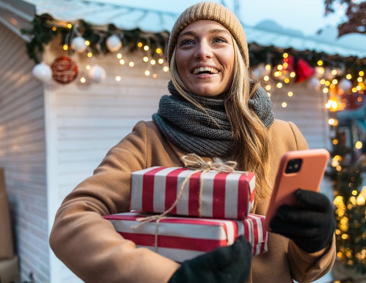 Junge Frau auf einem weihnachtlichen Markt, die lächelnd Geschenke trägt und ihr Smartphone benutzt