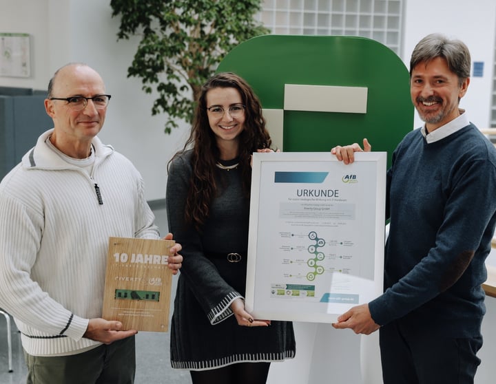 Drei Personen stehen in einem Innenraum und halten zwei Auszeichnungen. Die Person links hält eine Holzplakette zum zehnjährigen Jubiläum der Partnerschaft, während die beiden Personen in der Mitte und rechts eine gerahmte Urkunde präsentieren. Im Hintergrund ist ein großes grünes Riverty-Logo zu sehen.
