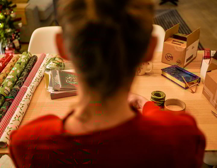 Eine junge Frau sitzt vor einem Tisch, der voll mit buntem Geschenkpapier und Weihnachtskarten ist