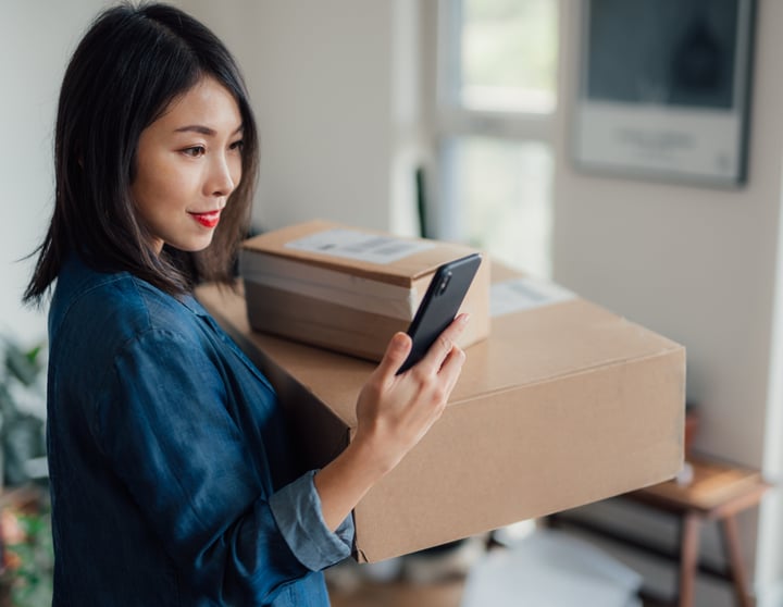 Eine Frau, die zwei Kartonschachteln in ihrem linken Arm trägt, schaut auf ihr Smartphone, während sie sich drinnen befindet. Sie trägt eine blaue Jacke und lächelt leicht. Im Hintergrund ist ein sanft beleuchteter Raum mit einem verschwommenen Schreibtisch und einem Fenster zu sehen.