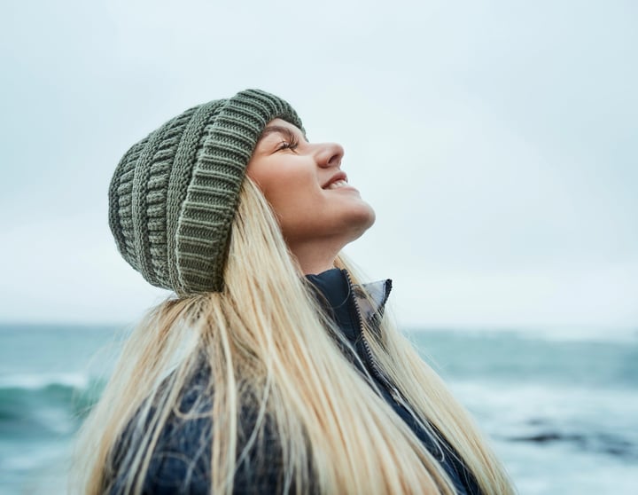Een jonge blonde vrouw heeft haar hoofd achterover en haar ogen gesloten en geniet van de koude bries aan zee