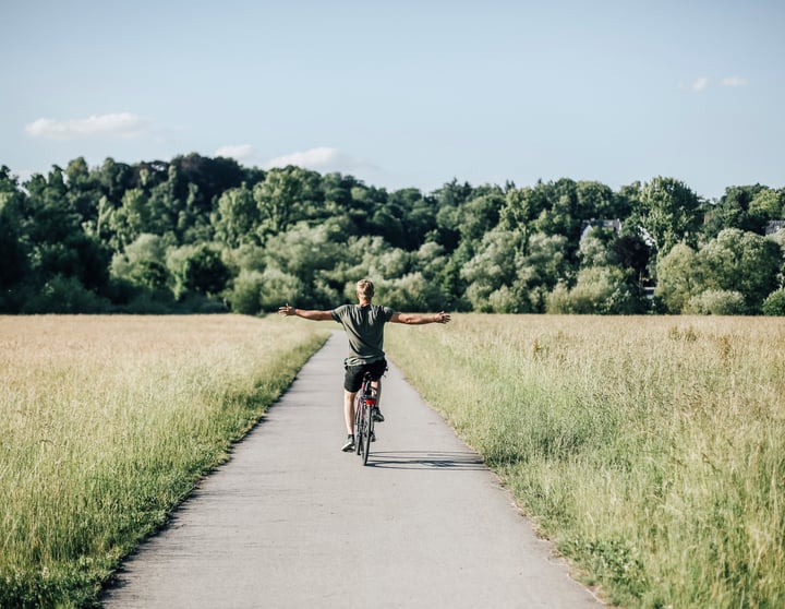 Ein Mann in einem grünen T-Shirt fährt mittig auf einer Straße Fahrrad und streckt dabei seine Arme von sich. Rechts und links von ihm befinden sich grüne Wiesen