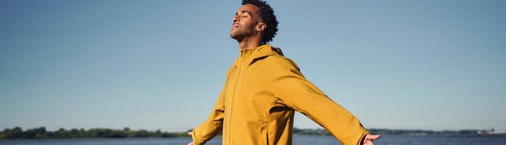Mann in gelber Jacke am Wasser mit ausgestreckten Armen, genießt den Moment.