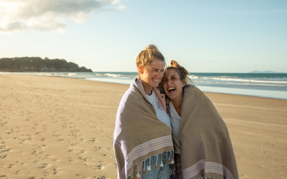 Twee jonge vrouwen lopen lachend over het strand met een deken om hun schouders