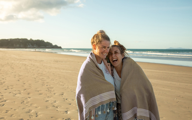 Twee jonge vrouwen lopen lachend over het strand met een deken om hun schouders