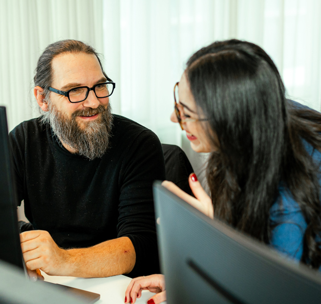 two riverty employees talking and laughing, sitting in front of a monitor