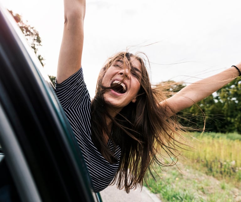 Junge Frau, die sich aus dem Fenster eines fahrenden Autos lehnt und dabei lacht