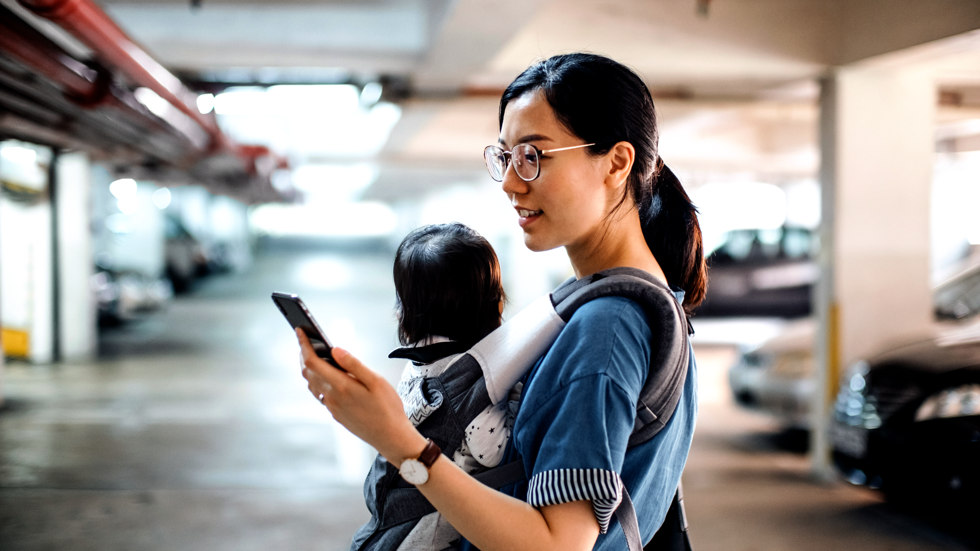 En kvinne som står i et parkeringshus med en baby på bæresele på magen, mens hun ser på telefonen sin