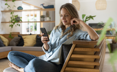 Eine Dame sitzt lächelnd auf der Couch und schaut auf ihr Telefon