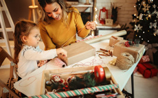 Eine Mutter und ihre Tochter verpacken gemeinsam Weihnachtsgeschenke während im Hintergrund ein geschmückter Weihnachtsbaum für festliche Stimmung sorgt.
