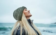 Eine junge blonde Frau hat ihren Kopf in den Nacken gelegt und ihre Augen verschlossen und genießt die kalte Brise am Meer